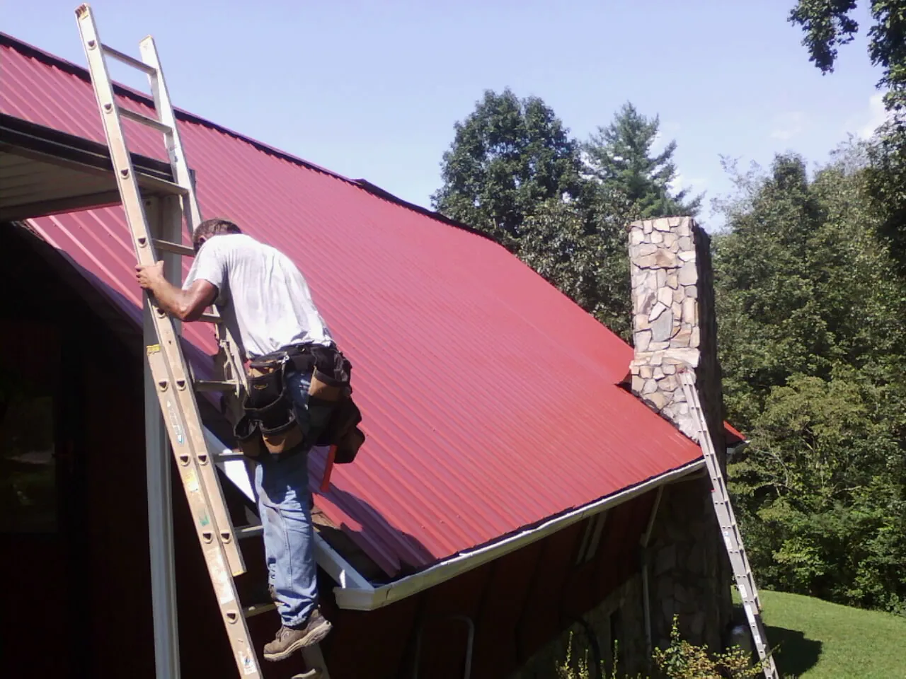 Roofer on a ladder beside a red metal roof and stone chimney — Soffit & Fascia Repair by Roofing Brothers in Valley Grove